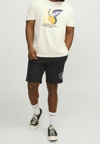 T-shirt crème avec imprimé graphique représentant un cercle jaune et une feuille bleue, associée à un short marine, des sneakers noires et des chaussettes blanches.