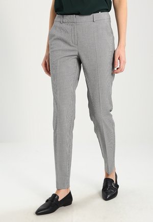 Femme portant un pantalon slim gris à carreaux avec passants de ceinture, associé à des mocassins noirs à bouts pointus et un haut vert foncé.