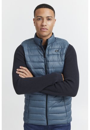 Dunkelblaue gepolsterte Weste mit Steppdesign und hohem Kragen, ausgestattet mit einem Reißverschluss auf der Vorderseite und marineblauen Strickärmeln, getragen über einem Marineblauen Shirt.