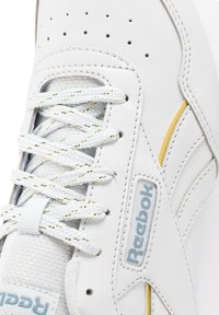 Zapatilla de cuero blanco con tejido gris texturizado, cordones trenzados en blanco y dorado, detalles perforados y un sutil acento amarillo en el lateral.