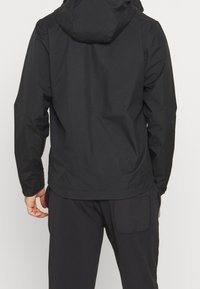 Mies, joka on pukeutunut mustaan huppariin ja mustiin housuihin, näkyy takaa yksinkertaista vaaleaa taustaa vasten.