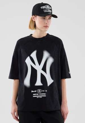 Mujer con una camiseta negra de gran tamaño de los New York Yankees y gorra a juego, mirando hacia un lado contra un fondo claro y liso.