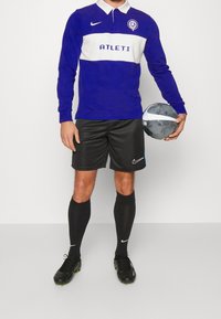 Lila långärmad polo med vit rand och texten "ATLETI", svarta sportshorts, svarta strumpor och svarta fotbollsskor, hållandes en svartvit fotboll.