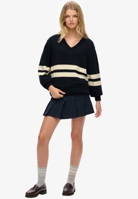 Granatowy sweter dzianinowy w kremowe paski, z dekoltem w kształcie litery V, połączony z granatową spódnicą w prążki. Modelka nosi szare i beżowe skarpetki w paski oraz mokasyny.