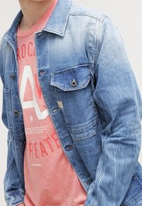 Jasnoniebieska kurtka dżinsowa z zapięciem na guziki, kieszeniami na piersiach i wyblakłym wyglądem. Nosi się ją na różowej koszulce z grafiką, na której znajduje się biały tekst.