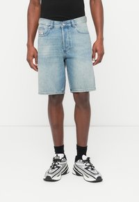 Shorts en denim bleu clair à coupe décontractée, avec cinq poches et une braguette à boutons. Portés avec des chaussettes noires montantes et des baskets sportives argentées et noires.