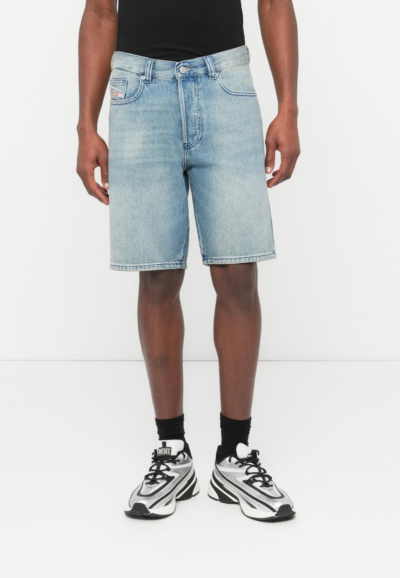 Shorts en denim bleu clair à coupe décontractée, avec cinq poches et une braguette à boutons. Portés avec des chaussettes noires montantes et des baskets sportives argentées et noires.