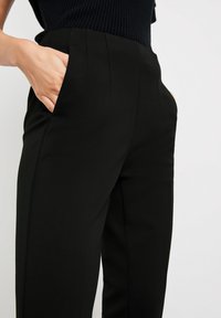 Pantalon noir sur mesure avec une texture lisse, doté de poches latérales et de plis à l'avant, présenté avec une main dans la poche.