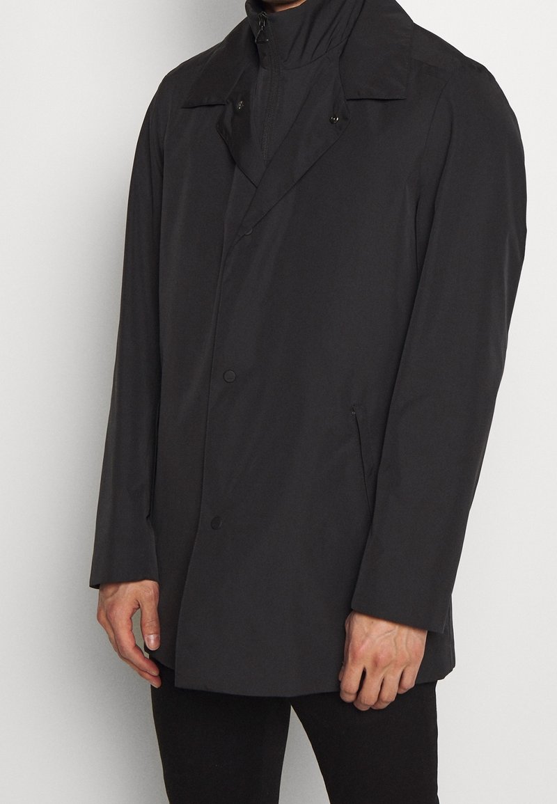 Czarny, średniej długości płaszcz z dwurzędowym zapięciem, wysokim kołnierzem i zamkiem błyskawicznym. Posiada boczne kieszenie i gładką fakturę.