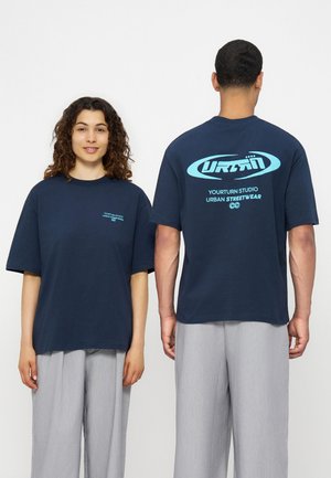 Femeie cu fața înainte și bărbat cu fața spre spate, purtând tricouri supradimensionate bleumarin cu text albastru deschis și pantaloni gri deschis, stând pe fundal alb.