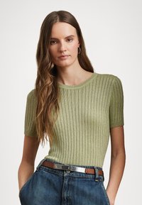 Short-sleeve ribbed top in light green, featuring a round neckline and textured knit. Paired with high-waisted denim jeans and a brown belt.