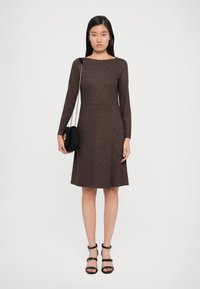Brown knit dress with long sleeves and a boat neckline, featuring a fitted bodice and flared skirt. Paired with a small black bag and strappy heels.