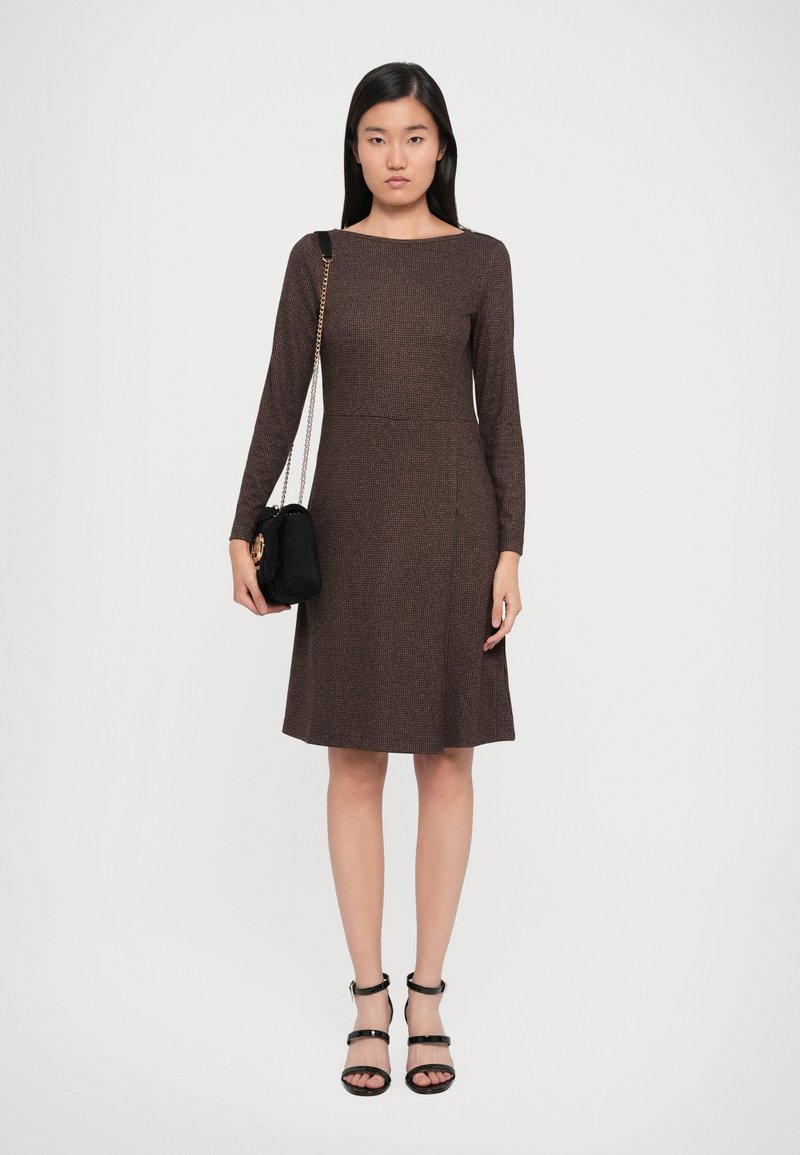 Brown knit dress with long sleeves and a boat neckline, featuring a fitted bodice and flared skirt. Paired with a small black bag and strappy heels.