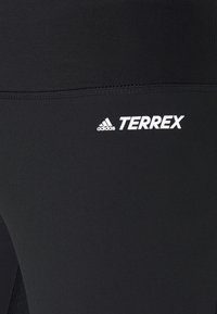 Leggings atletici neri con una texture liscia. Presentano i loghi bianchi "adidas" e "TERREX" sul lato sinistro inferiore.