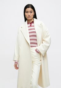 Pitkä kerma-valkoinen villakangastakki, jossa on lovettu kaulus, sivutaskut ja pehmeä tekstuuri, kerroksittain päällekkäin puetun vaaleanpunaisen ja tummanpunaruskean raidallisen neulepuseron sekä valkoisten housujen kanssa.