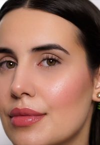 Primer plano de un rostro con piel suave y radiante, ojos marrón claro y labios llenos que llevan un lápiz labial nude brillante. Se pueden ver pendientes de aro dorados.