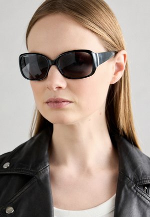 Zwarte oversized zonnebril met een glanzende afwerking, voorzien van een gedurfde frameontwerp en donkere lenzen, geplaatst tegen een neutrale achtergrond.