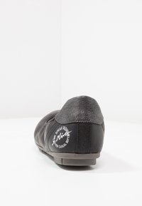 Black casual shoe with textured leather, rounded shape, and a low heel. Features a metallic-accented back with a logo print on the side.