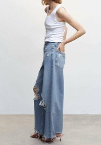 Jean large bleu clair avec des déchirures usées, taille haute et coupe décontractée. Porté avec un débardeur blanc ajusté et des sandales roses.