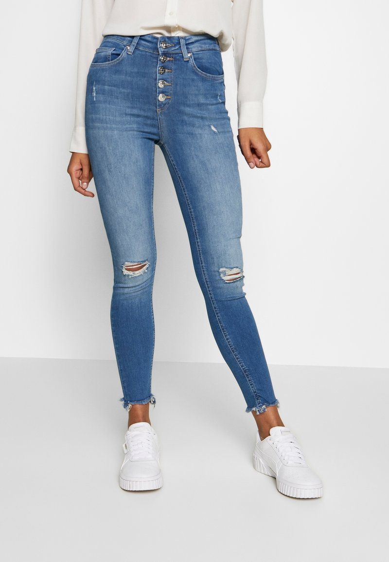 Jeans de denim azul de cintura alta con cierre de botón, detalles desgastados y dobladillo deshilachado. Llevados con zapatillas blancas.