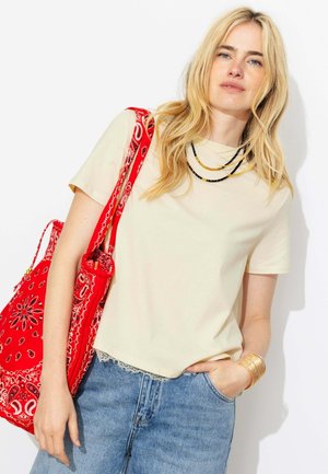 Femme aux longs cheveux blonds portant un t-shirt crème, des colliers de perles superposés, un jean bleu, un bracelet manchette doré, et portant un sac fourre-tout rouge à motifs.