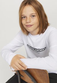 Biała koszula z długim rękawem i czarnym tekstem logo, noszona przez osobę opierającą ramiona na drewnianym krześle, o gładkiej fakturze i prostym designie.