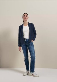 Chaqueta de estilo navy con cremallera sobre una camiseta blanca, combinada con unos vaqueros azules y zapatillas blancas. Diseño sencillo, tejido suave y estilo informal.