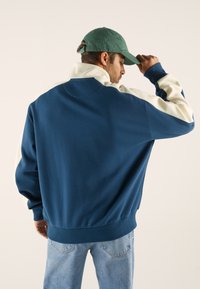 Pullover blu e crema con colletto alto, caratterizzato da una texture morbida e orlo a coste. Indossato con jeans azzurri chiari e un cappellino verde.
