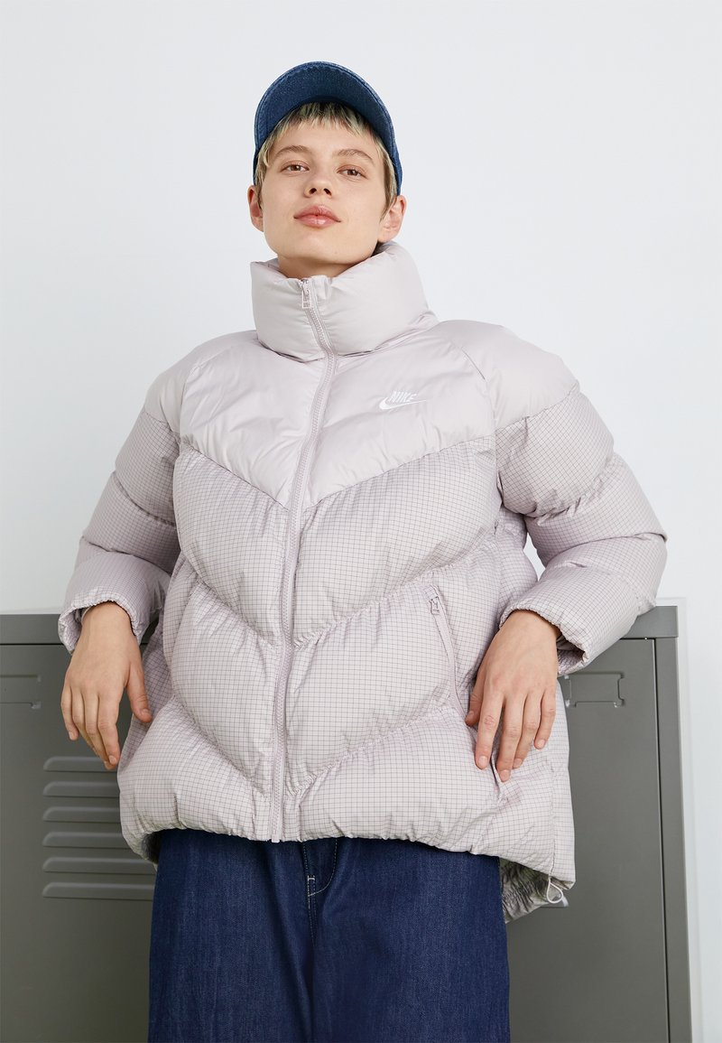 Doudoune violette clair avec un col haut, présentant un motif géométrique, une fermeture éclair et le logo Nike. Portée sur un denim bleu.