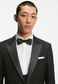 Giacca da smoking nera con rever in raso, camicia bianca e papillon nero. Presenta un fazzoletto da taschino e un tessuto testurizzato.