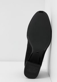 Chaussure à talon haut en daim noir avec un bout lisse et une semelle texturée, dotée d'un talon recouvert et d'un logo sur le dessous.