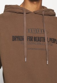 Sudadera de algodón marrón con cordones, que presenta texto impreso en azul marino oscuro. Textura suave y corte relajado. Detalles destacados del logo.