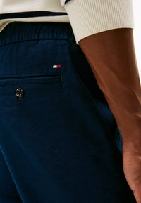 Námorně modré lněné šortky s kapsou na zadní straně opatřenou knoflíkem, zdobené malou pruhovanou výšivkou v červené, bílé a modré barvě na boku.