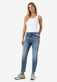 Hvit ribbestrikket tanktopp, lyseblå høytlivsjeans og sølvfargede sportssko. Modellen står med hendene i lommene foran en ensfarget bakgrunn.