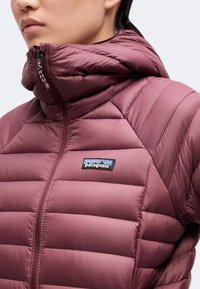 Personne portant une doudoune bordeaux Patagonia avec un col montant zippé et un logo visible sur la poitrine.