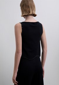 Top negro sin mangas hecho de tela de punto, con dobladillo en forma de conchas y costuras adornadas, visto desde atrás.