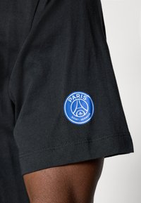 T-shirt en coton noir avec un patch circulaire bleu portant les textes "PARIS" et "SAINT-GERMAIN", ainsi qu'un design de logo sur la manche gauche.