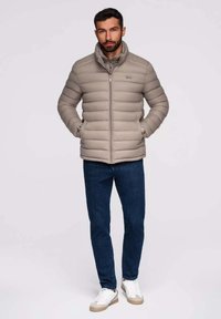 Beige donsjas met horizontaal gestikt patroon, ritssluiting en staande kraag. Gecombineerd met donkere jeans en lichte sneakers.
