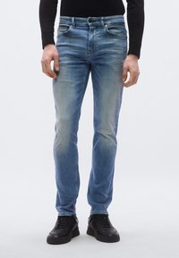 Svetlomodré džínsové nohavice s úzkym strihom, s efektom opotrebovania, s piatimi vreckami a zapínaním na gombík. Nosia sa s čiernymi teniskami.