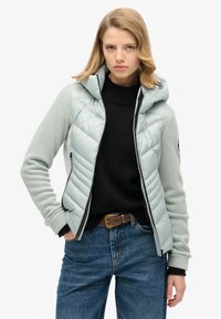 Chaqueta aislante gris claro con un diseño acolchado, cierre de cremallera y capucha. Presenta mangas en contraste y un suéter de punto negro debajo.