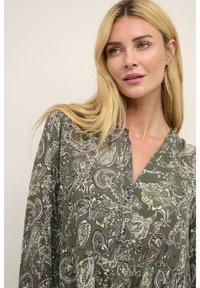 Rochie lungă de culoare verde, cu un imprimeu paisley în alb și verde deschis, având un decolteu în V și detalii cu nasturi pe față. Textură moale.