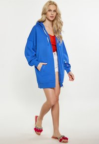 Felpa blu over-size con zip frontale, cappuccio con cordino e tasche. Indossata sopra una maglietta rossa e pantaloni corti bianchi, con accenti colorati.
