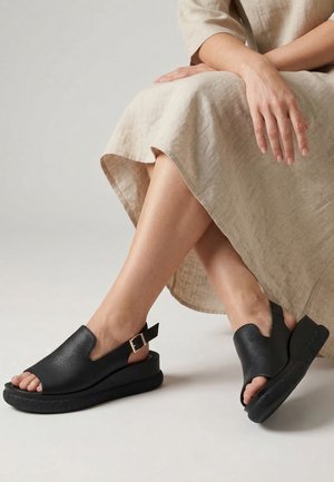 Person iført en beige kjole siddende med krydsede ben, der viser sorte åbne lædersandaler med spænder og tykke såler.