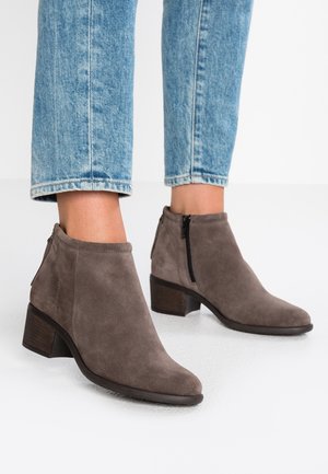 Personne portant des bottines en suédine grise avec des fermetures éclair latérales et des talons blocs, associées à un jean en denim bleu clair coupé au-dessus des chevilles.