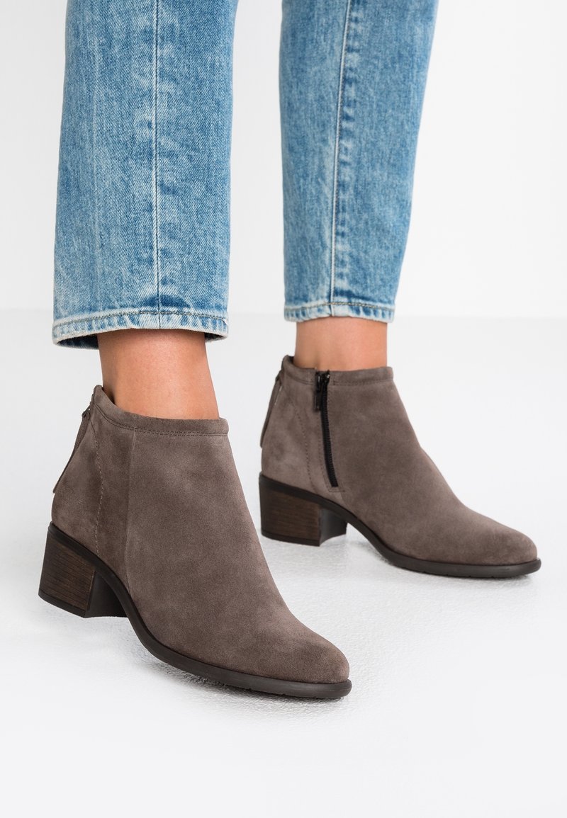 Personne portant des bottines en suédine grise avec des fermetures éclair latérales et des talons blocs, associées à un jean en denim bleu clair coupé au-dessus des chevilles.