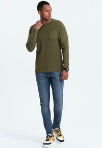 Oliivinvihreä pitkähihaine paita, jossa on ribbikuviointi ja rintatasku, yhdistettynä istuviin sinisiin farkkuihin ja monivärisiin tennareihin.