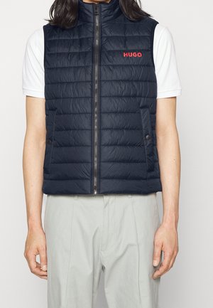 Mężczyzna noszący granatową pikowaną kamizelkę z przednim zamkiem błyskawicznym i czerwonym logo "HUGO", białą koszulę z krótkim rękawem oraz jasnoszare spodnie.