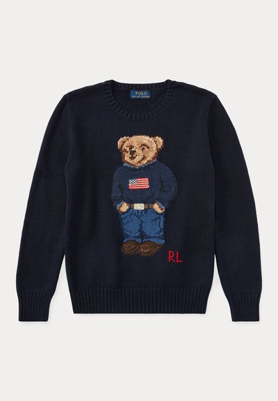 Pull bleu marine avec un ours en peluche tricoté portant un jean, des chaussures marron et un pull bleu marine avec un drapeau américain sur le devant et les initiales rouges "RL".