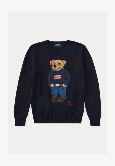 Pull bleu marine avec un ours en peluche tricoté portant un jean, des chaussures marron et un pull bleu marine avec un drapeau américain sur le devant et les initiales rouges "RL".