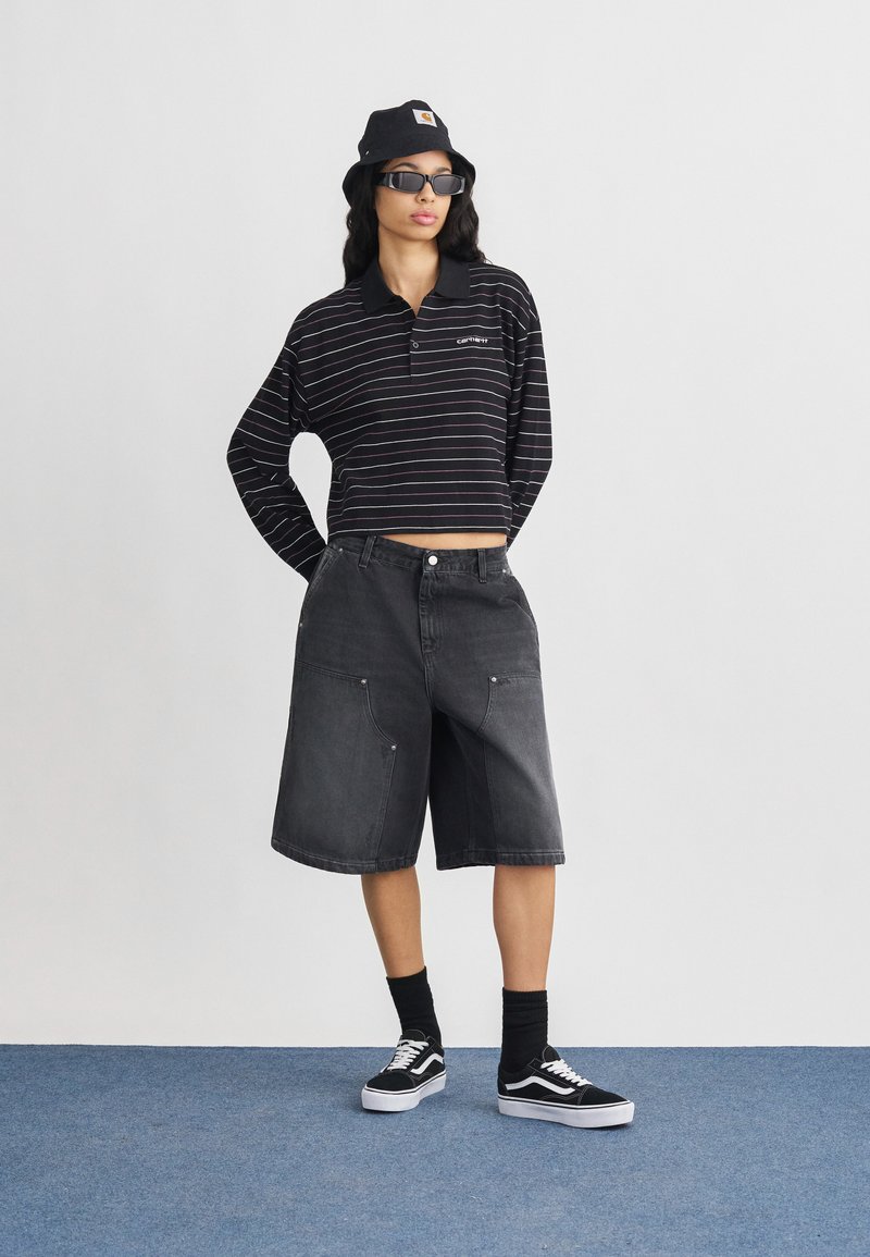 Vrouw met een zwarte bucket hat, zonnebril, gestreepte cropped top met lange mouwen, zwarte knielange shorts, zwarte sokken en zwarte Vans sneakers.